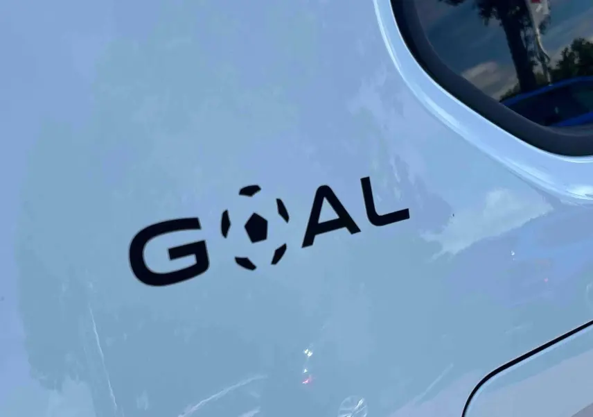 Détail du logo "GOAL" noir sur la porte arrière blanche d’un Volkswagen Touran 2025, vue latérale droite.
