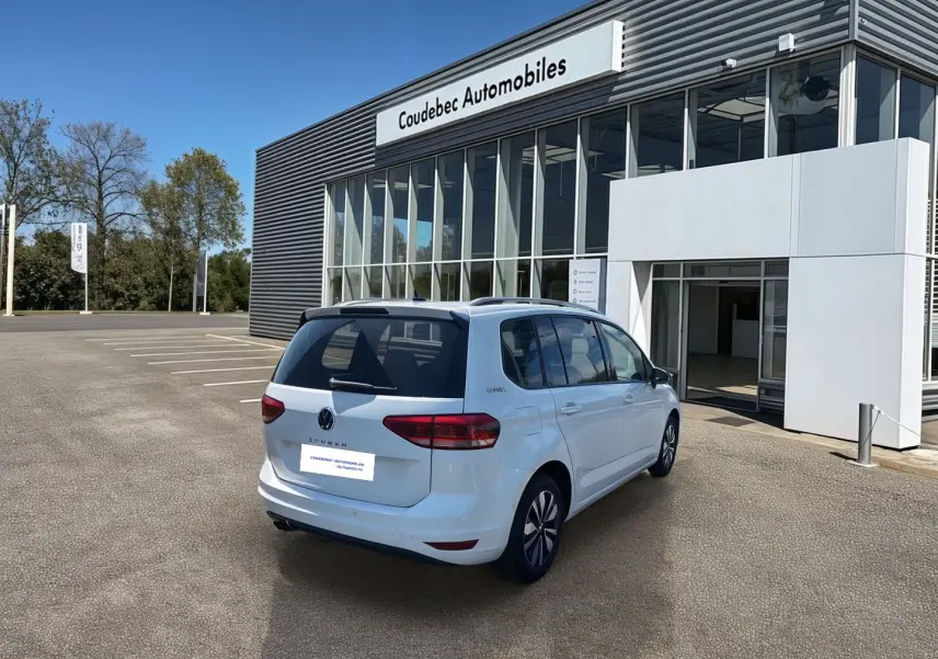 Volkswagen Touran blanc vu en 3/4 arrière droit devant un bâtiment de concession Coudebec Automobiles.