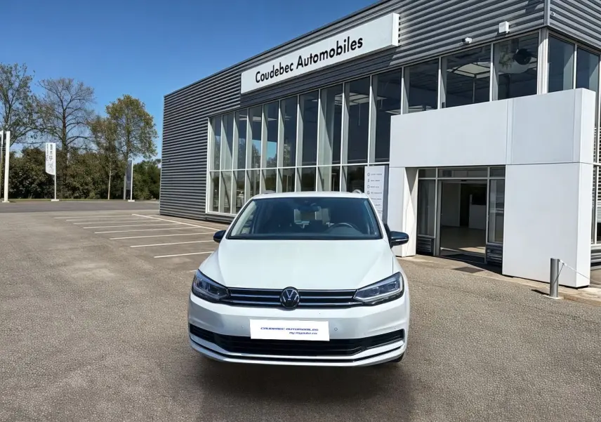 Volkswagen Touran blanc vue de face, stationné devant un bâtiment commercial sous un ciel bleu.