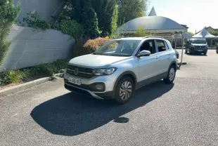 Volkswagen T-Cross gris clair en 3/4 avant droit, stationné sur un parking ensoleillé avec végétation en arrière-plan.