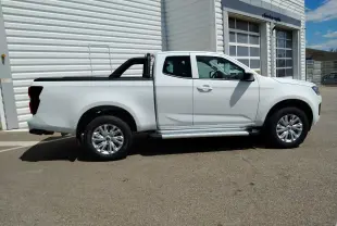 Vue de profil côté droit d'un pick-up Isuzu D-MAX blanc avec arceau noir et couvre-benne roulant, stationné devant un bâtiment industriel.