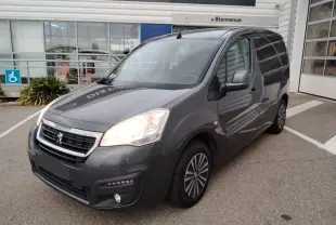 Peugeot Partner Fourgon gris platinium en 3/4 avant droit avec phares allumés, stationné devant un bâtiment commercial.