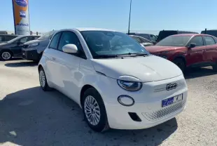 Vue 3/4 avant d'une FIAT 500 électrique blanche avec logo distinctif et plaque "Zero KM" sur un parking extérieur.