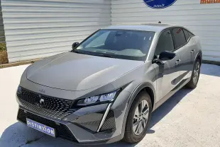 Peugeot 408 gris selenium vue 3/4 avant droit, avec calandre distinctive et phares LED anguleux, stationnée devant un mur blanc.