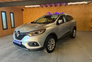 Renault Kadjar gris en 3/4 avant droit dans un garage avec éclairage intérieur et logo néon bleu en arrière-plan.