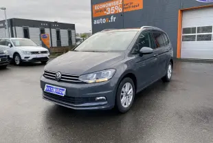 Volkswagen Touran gris dauphin en 3/4 avant droit sur parking devant un bâtiment commercial.