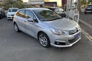 Citroën C4 gris clair vue 3/4 avant droit, stationnée devant un restaurant avec reflets métallisés.
