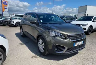 Peugeot 5008 gris clair vue 3/4 avant droit sur parking avec ciel partiellement nuageux.