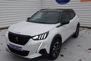 Peugeot 2008 blanc nacré avec toit noir en 3/4 avant droit, phares allumés et attelage visible à l'arrière.