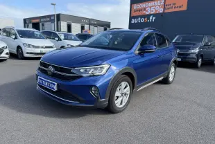 Volkswagen Taigo bleu récif métallisé vu en 3/4 avant droit sur parking devant un panneau publicitaire orange.