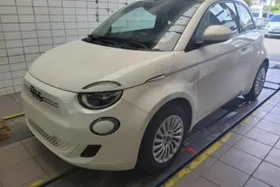 Vue 3/4 avant droite d'une Fiat 500 électrique blanche stationnée dans un garage avec phares ronds distinctifs.