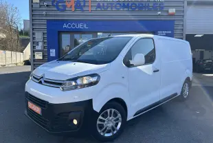 Vue 3/4 avant droite d'un Citroën Jumpy blanc avec attelage, garé devant un bâtiment commercial.