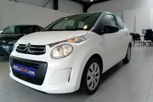 Citroën C1 blanche vue 3/4 avant droit en intérieur avec phares allumés et enjoliveurs noirs sur jantes.