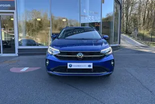 Volkswagen Taigo bleu Recife vue de face, avec calandre noire et phares LED allumés devant showroom.