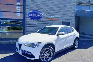 Alfa Romeo Stelvio blanc en 3/4 avant droit, stationné devant un bâtiment avec logo Distinxion visible.