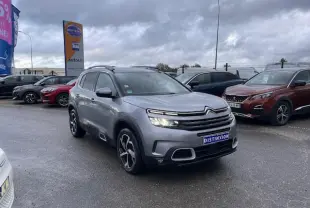 Citroën C5 Aircross gris Artense vue 3/4 avant avec feux allumés sur parking sous ciel nuageux.