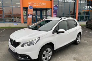 Peugeot 2008 blanc vue 3/4 avant droit, avec jantes acier et barres de toit, stationné devant un garage orange.