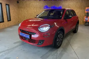 Vue 3/4 avant d'une FIAT 600 rouge 2024 avec phares allumés, calandre noire et jantes stylisées en intérieur bois.