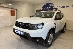 Dacia Duster blanc en 3/4 avant droit, avec calandre noire et barres de toit, présenté en intérieur showroom.