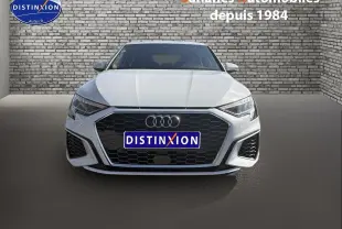 Vue frontale d'une Audi A3 Sportback blanche 2023 avec calandre noire et projecteurs LED, sur fond mural en briques.