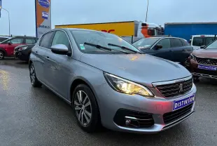 Peugeot 308 gris Artense vue 3/4 avant droit sous la pluie avec phares allumés sur parking extérieur.