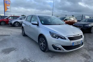 Peugeot 308 gris aluminium vue 3/4 avant droit, avec phares LED et jantes alliage sur parking extérieur.