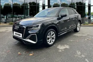Audi Q2 noir vue 3/4 avant droit, avec calandre distinctive et jantes argentées sur parking devant vitres réfléchissantes.