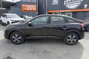 Profil droit d'une Citroën C4 électrique noire 2025 avec inserts noir brillant, stationnée devant un garage.