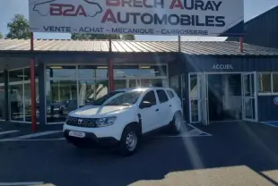 Dacia Duster blanc en 3/4 avant droit stationné devant un garage Brech Auray Automobiles avec portes vitrées ouvertes.