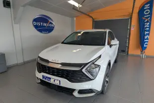 Vue 3/4 avant d'un KIA Sportage blanc 2025 avec calandre noire, jantes alliage bicolores et barres de toit visibles en intérieur showroom.