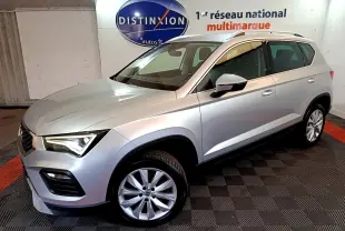 Vue 3/4 avant droit d’un SEAT Ateca gris clair 2022, avec jantes alliage et barres de toit chromées, en intérieur showroom.