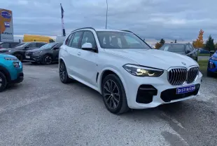 BMW X5 xDrive 30d blanc en 3/4 avant droit avec calandre M Sport et phares allumés sur parking extérieur.