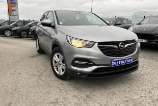 Opel Grandland X gris clair vu en 3/4 avant droit avec phare allumé et jantes alliage 17 pouces sur parking.