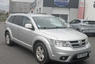 Fiat Freemont gris en 3/4 avant droit, avec barres de toit et vitres arrière surteintées, stationné devant un garage.