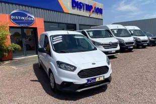 Ford Tourneo Courier blanc vue 3/4 avant droit, fourgon utilitaire devant un garage avec plusieurs véhicules blancs alignés.