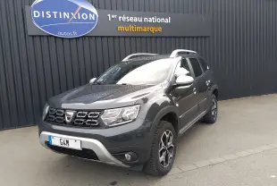 Dacia Duster gris 3/4 avant droit avec jantes alliage et barres de toit, devant un mur noir avec enseigne Distinxion.
