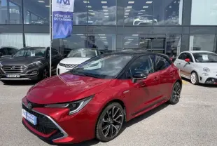 Toyota Corolla 184 H Collection rouge métal avec toit noir, vue 3/4 avant droit, garée devant un showroom.