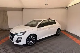 Peugeot 208 blanche vue en 3/4 avant droit, avec jantes noires et phares LED allumés, stationnée en intérieur.