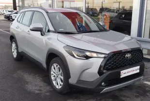 Toyota Corolla Cross gris acier vue 3/4 avant droit, avec calandre noire et phares allumés, stationnée devant un garage.