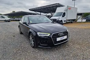 Audi A3 Sportback noire vue 3/4 avant droit sur un sol gravillonné avec des véhicules utilitaires en arrière-plan.