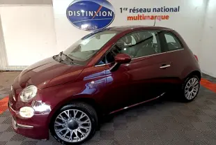Vue 3/4 avant gauche d'une FIAT 500 rouge bordeaux avec jantes alliage et phares ronds allumés en intérieur showroom.