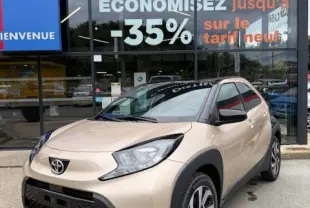 Toyota Aygo X beige vue 3/4 avant droit, avec jantes alliage et détails noirs contrastants sur carrosserie compacte.