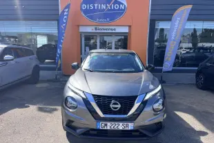 Nissan Juke 2023 gris argile vue de face devant une concession avec drapeaux publicitaires.