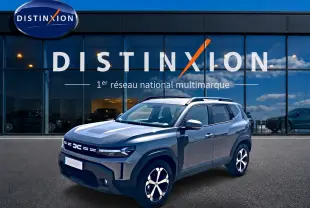 Vue 3/4 avant droit du Dacia Duster gris schiste avec jantes alu et barres de toit devant une vitrine Distinxion.