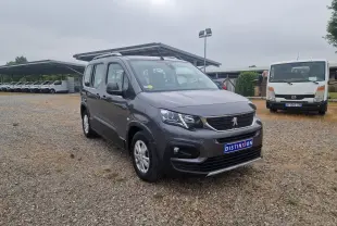 Peugeot Rifter gris platinium en 3/4 avant droit sur un sol gravillonné avec plusieurs véhicules en arrière-plan.