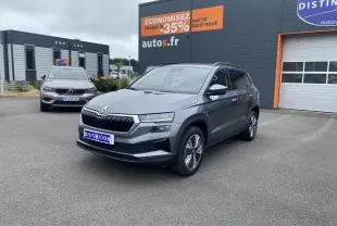 SUV Skoda Karoq gris graphite métallisé vu en 3/4 avant droit devant un garage avec un autre SUV en arrière-plan.