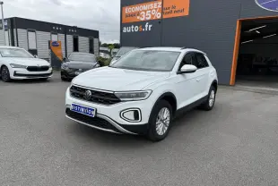 Volkswagen T-Roc blanc pur vu en 3/4 avant droit sur parking devant un garage avec panneau promotionnel orange.