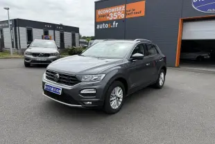 Volkswagen T-Roc gris Indium Metal en 3/4 avant droit, stationné devant un garage avec panneaux publicitaires.
