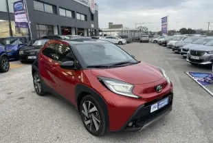 Toyota Aygo X rouge piment avec toit noir, vue 3/4 avant droit, stationnée devant un garage en extérieur.