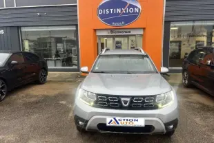 Vue frontale d'un Dacia Duster 1.5 Blue dCi gris Comète métallisé avec barres de toit, phares allumés, devant un garage.
